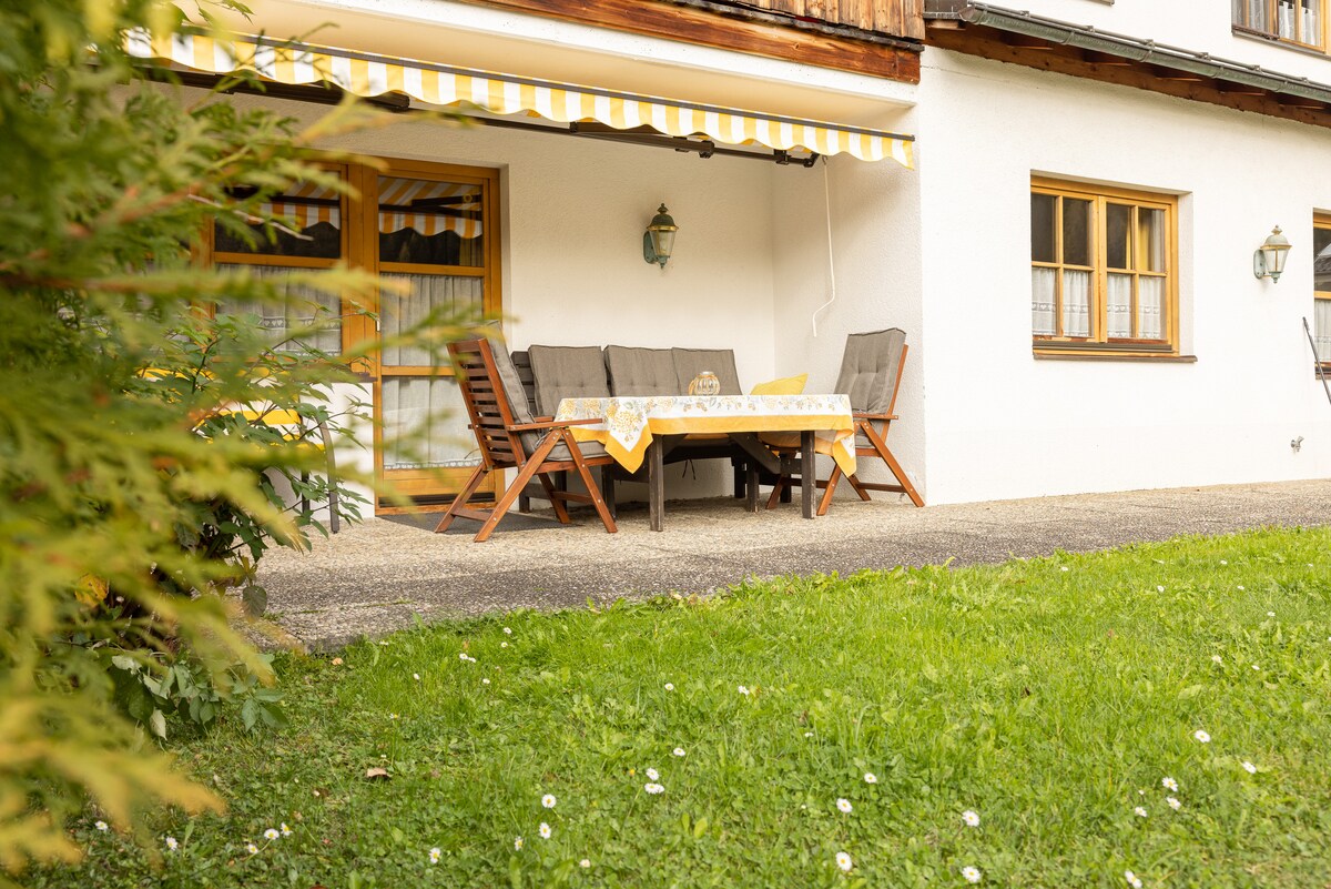 An outdoor seating area is featured under a striped awning, with a wooden table surrounded by comfortable chairs. The open space is complemented by a well-maintained lawn, dotted with small flowers, creating a serene environment for relaxation.