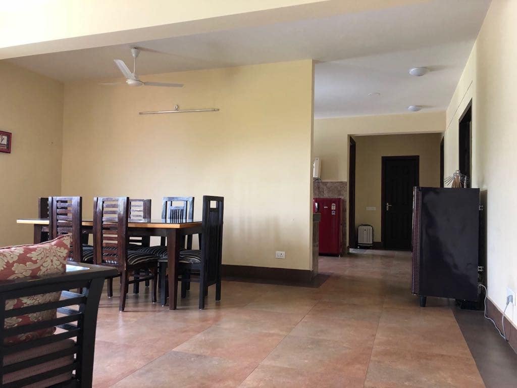 A spacious dining area features a large wooden table surrounded by six chairs. A ceiling fan is visible, while a refrigerator and a cabinet are situated in the background. The flooring consists of neutral-toned tiles, contributing to the uncluttered ambiance.