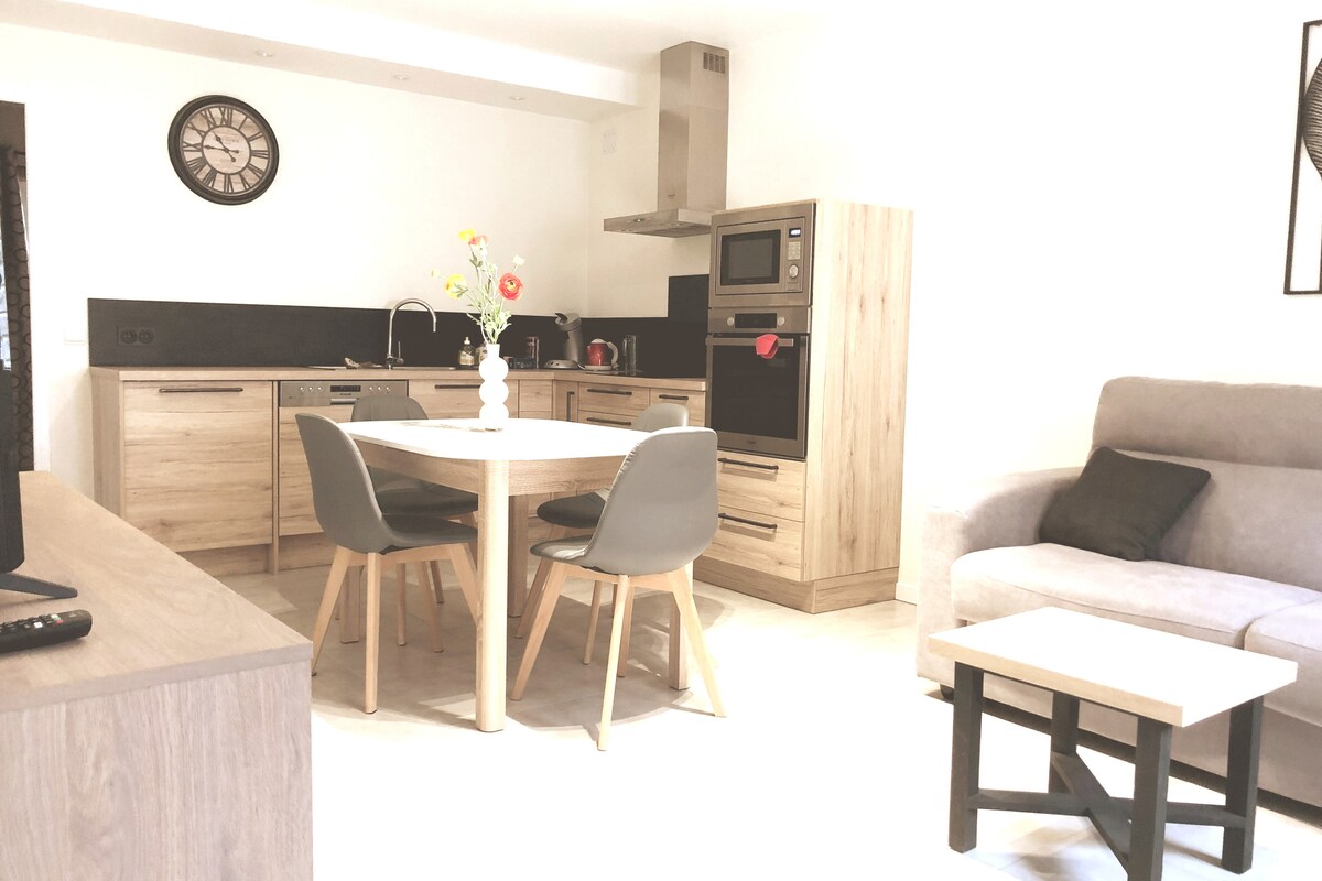 A modern kitchen and dining area are displayed, featuring wooden cabinetry and black countertops. A round dining table with four chairs is positioned in the center. Appliances, including a microwave and oven, are integrated into the design. A gray sofa adds comfort to the space.