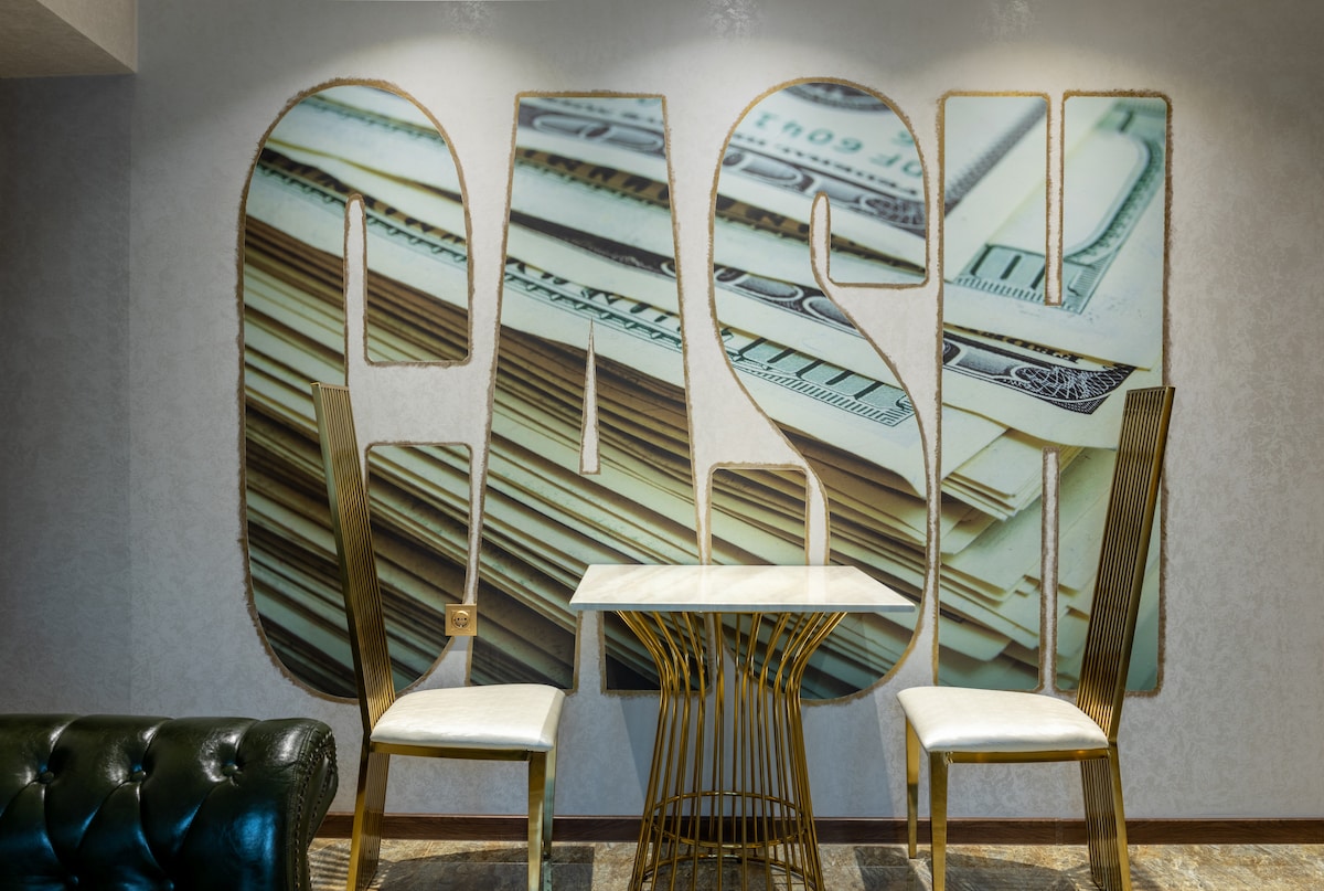 A modern dining space is showcased with two elegant chairs positioned around a sleek, round table. Behind the table, a prominent wall graphic depicts stacks of cash, providing a unique artistic element to the room.