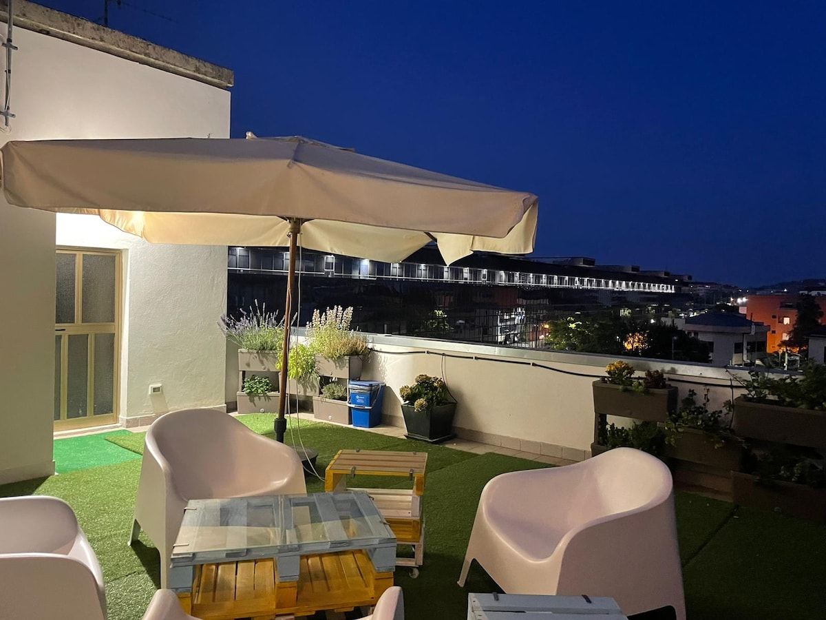 An outdoor terrace is shown at night, featuring several white chairs and a glass-top table. A large umbrella provides shade. The space is accented with potted plants, creating a cozy environment for relaxation.