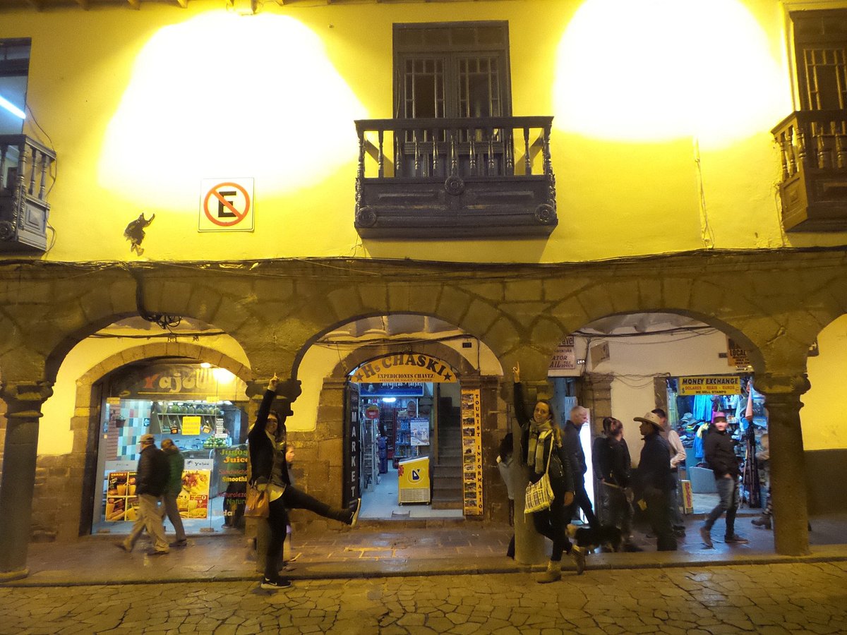 A lively street scene is depicted, showcasing arched stone walkways and illuminated storefronts. The warm overhead lighting highlights the vibrant activity of passersby. Balconies above the shops display ornate wooden railings, adding to the character of the building facade.