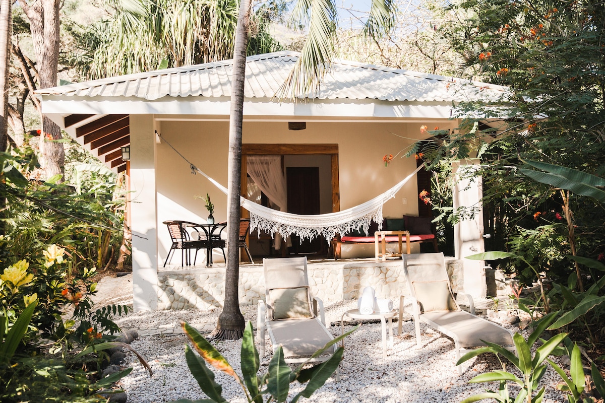 A charming bungalow is surrounded by lush greenery, featuring a hammock delicately hung between two posts. A small patio with a table and chairs invites relaxation, while sun loungers are positioned in the foreground, offering a serene spot to enjoy the natural setting.