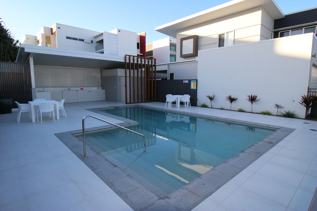 A modern pool area features a rectangular swimming pool surrounded by a tiled deck. White outdoor furniture, including tables and chairs, is positioned nearby. The area is enclosed by contemporary architectural elements, with tropical plants adding a touch of greenery.