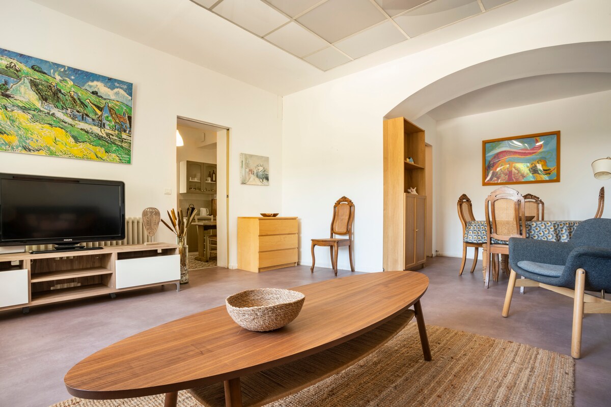 A spacious living area features a wooden coffee table centered on a soft rug. The room is complemented by artwork on the walls, a television unit, and a dining table with six chairs. Natural light is filtered through the ceiling, enhancing the neutral color palette.