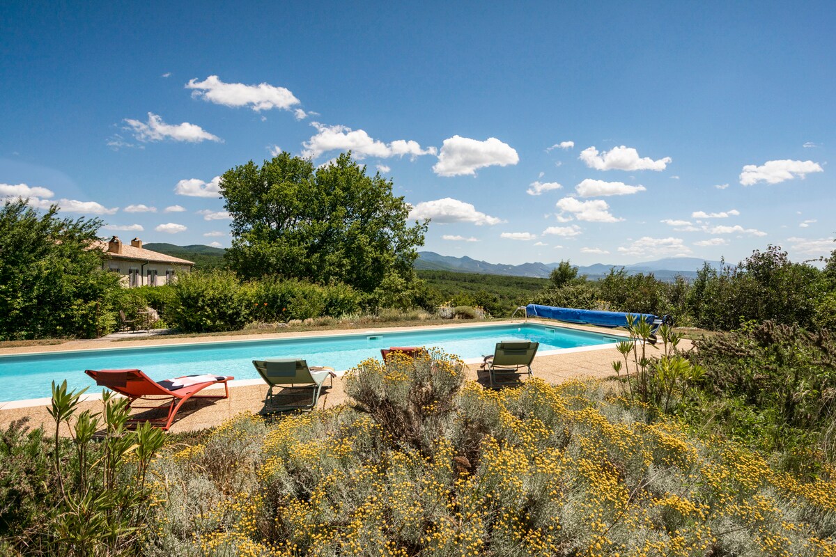A serene view of a heated swimming pool surrounded by lush greenery and wildflowers. Comfortable lounge chairs are positioned nearby, offering a perfect spot for relaxation. The backdrop features rolling hills under a bright blue sky, enhancing the peaceful outdoor environment.