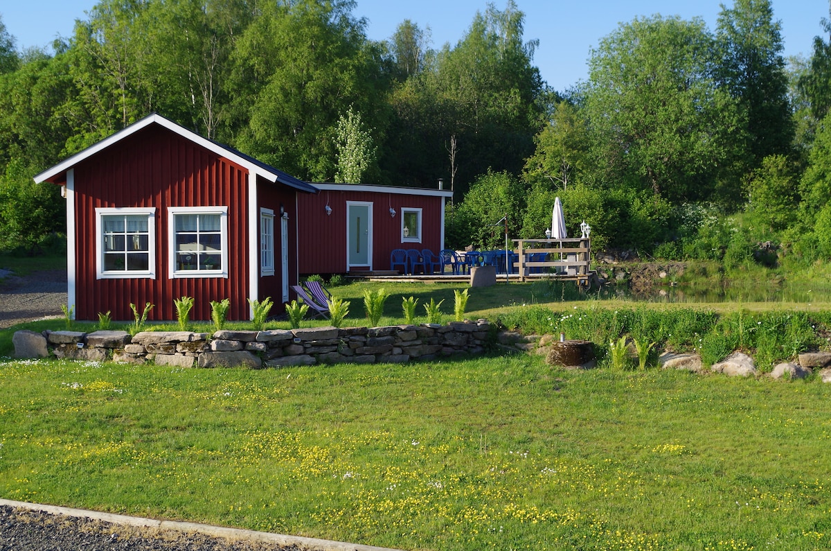 A charming red cottage is surrounded by lush greenery, with large windows providing natural light. The yard features a stone border, vibrant wildflowers, and a terraced area with outdoor seating, creating a serene atmosphere near a small pond.