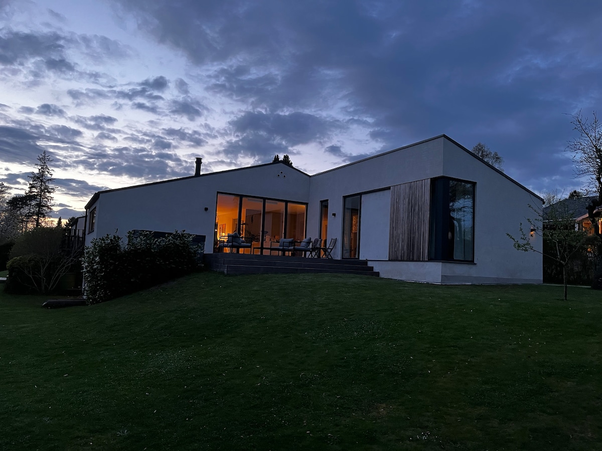 The modern villa is set against a twilight sky, featuring expansive glass doors that reveal warm interior lighting. A spacious lawn surrounds the home, with a subtle slope leading to the entrance. Outdoor seating is visible on the deck, inviting relaxation.
