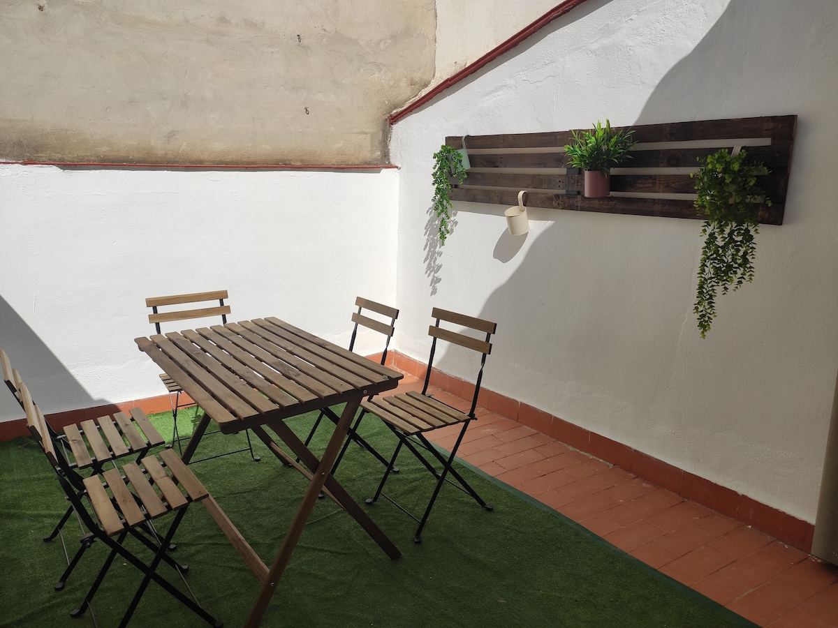 An outdoor terrace features a wooden table surrounded by four folding chairs. Against the white wall, wooden shelving displays potted plants, contributing to a fresh ambiance. A green carpet covers the floor, enhancing the space's inviting atmosphere for outdoor dining.