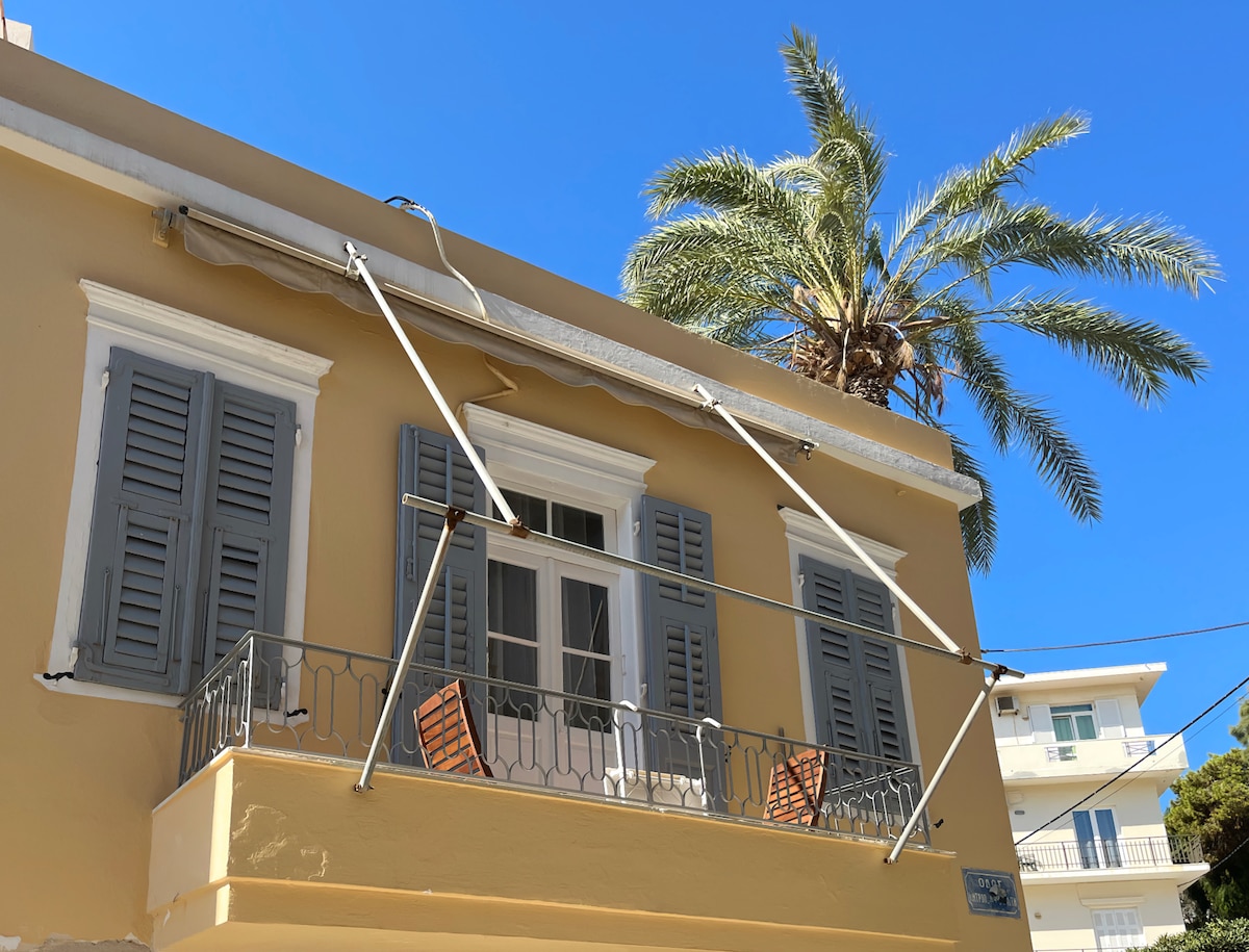 The exterior of a corner house is shown, featuring light yellow walls and grey shutters. A balcony is visible with two wooden chairs, alongside a railing adorned with decorative elements. A palm tree stands nearby against a clear blue sky.