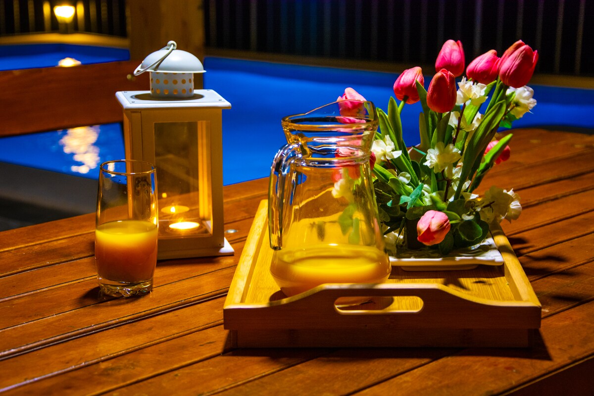 A wooden table is set with a glass pitcher and a glass containing a refreshing beverage. A tray holds a small floral arrangement of pink and white flowers. Nearby, a lantern emits a soft glow against the backdrop of a softly lit pool.