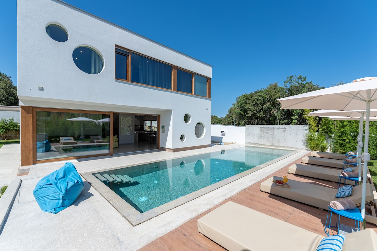 A modern villa features a large outdoor pool surrounded by lounge chairs. The exterior showcases circular windows and a mix of white and natural wood finishes. An inviting seating area is visible through expansive glass doors, blending indoor and outdoor spaces seamlessly.