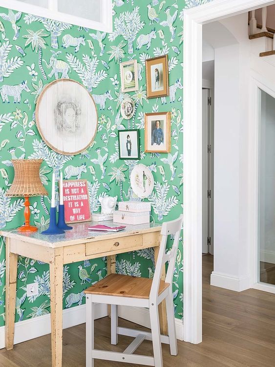 A small desk is set against a vibrant green patterned wallpaper featuring botanical and animal motifs. Above the desk, a variety of framed pictures add visual interest. A wooden chair is positioned at the desk, while light-colored flooring contributes to a bright atmosphere.
