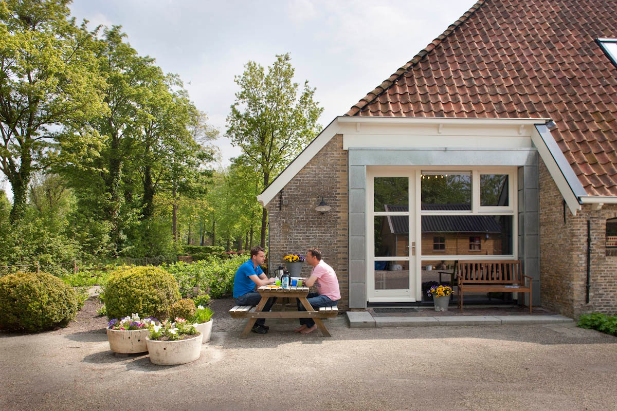 An outdoor seating area is shown with a picnic table and chairs surrounded by potted flowers. In the background, greenery and trees are visible, along with the exterior of a modern building featuring large glass doors and a sloped roof.