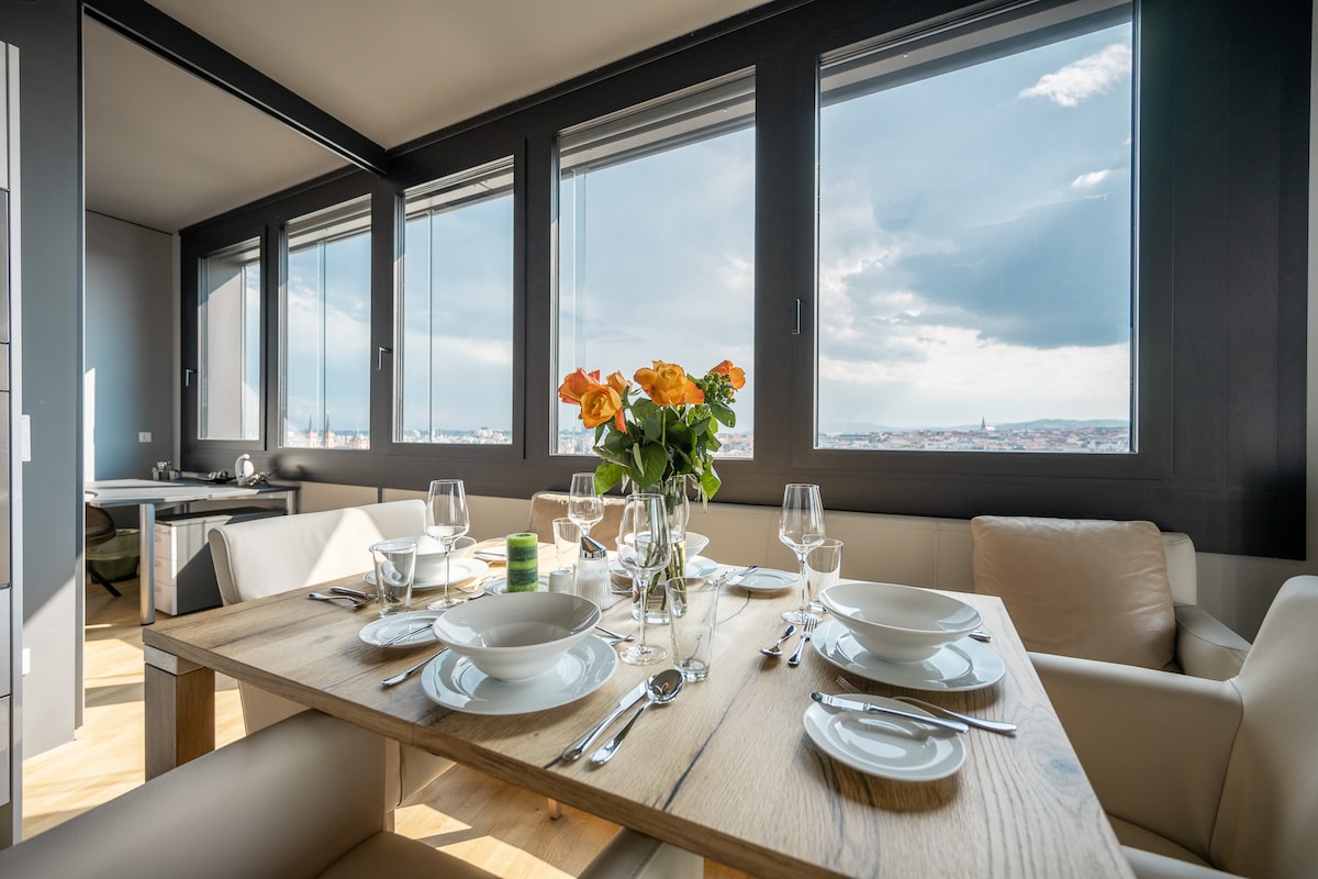 A dining area is presented with a wooden table set for four. Elegant glassware and utensils are arranged neatly beside white plates. A vase of fresh orange roses adds a pop of color, while large windows frame a view of the city skyline and bright sky.