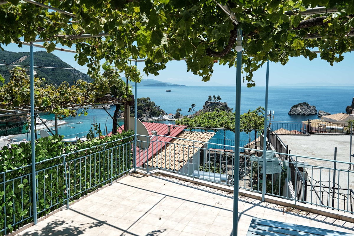 The shared roof terrace offers a scenic view of the Ionian Sea and surrounding islands, framed by lush green vines overhead. A tiled floor extends across the terrace, enhancing the spaciousness of the area, ideal for relaxation or dining while enjoying the ocean vista.