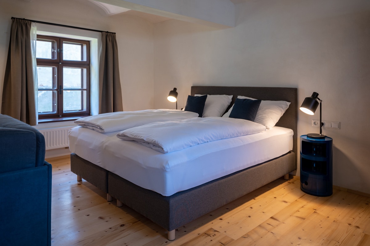 A spacious sleeping area features a large, comfortable bed with multiple pillows. Soft lighting is provided by bedside lamps on either side. Natural light enters through a window adorned with curtains, highlighting the warm wooden flooring throughout the room.