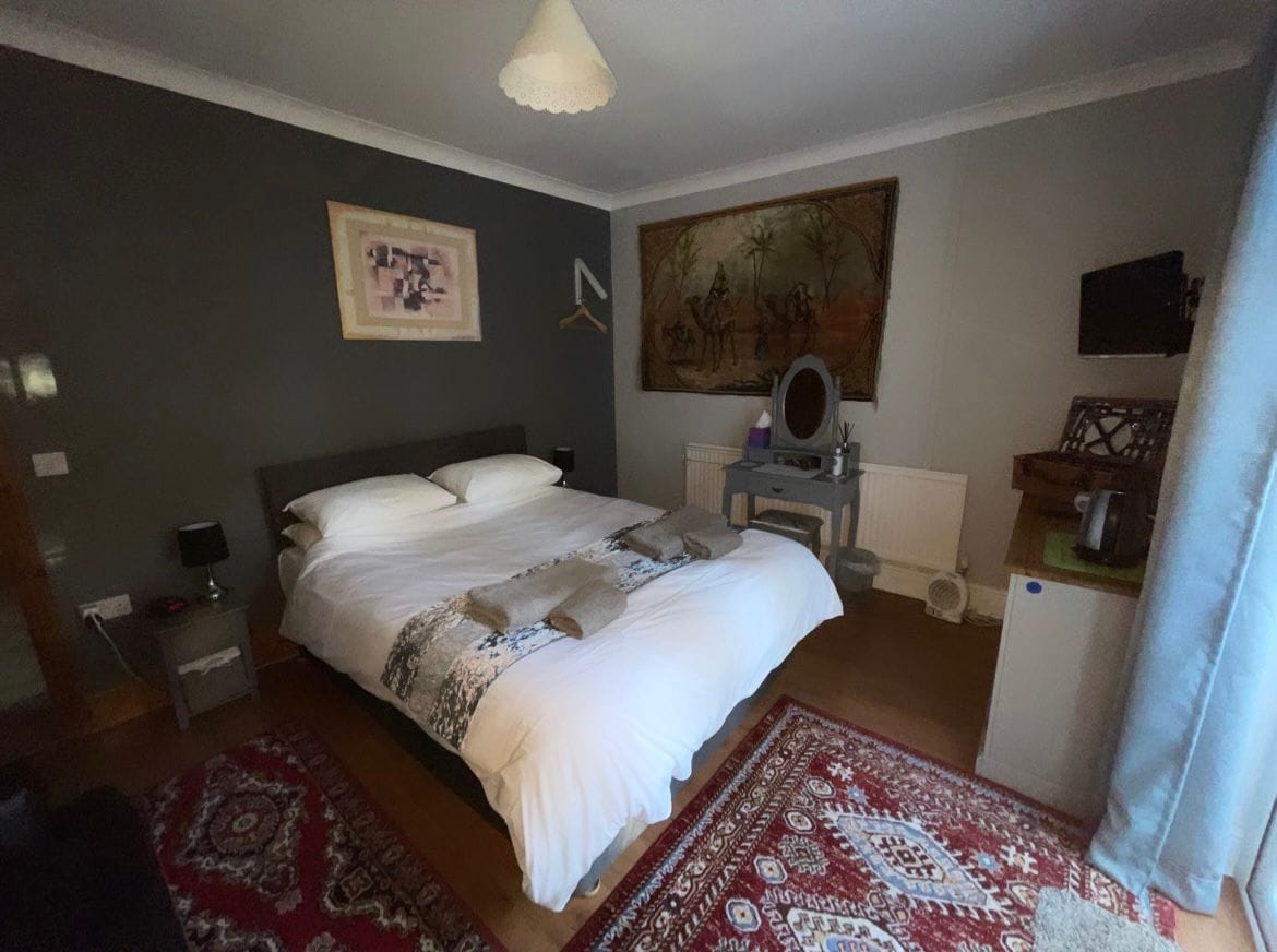 The room features a spacious king-size bed dressed in crisp linens, accompanied by bedside tables. A wall-mounted TV is positioned opposite a dressing table, with a decorative rug adding warmth to the wooden floor. Natural light filters in through a window with sheer curtains.