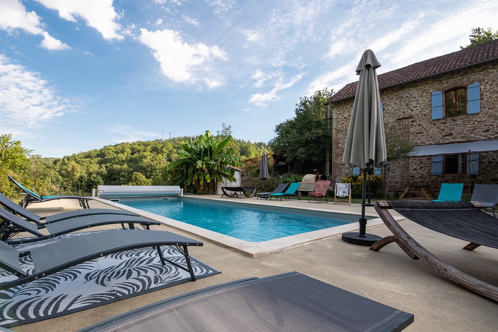 A tranquil outdoor area features a salt-water pool surrounded by a spacious deck and sun loungers. Lush greenery and trees offer a natural backdrop, while a large umbrella stands beside the pool, providing shade. The stone building is seen in the background.
