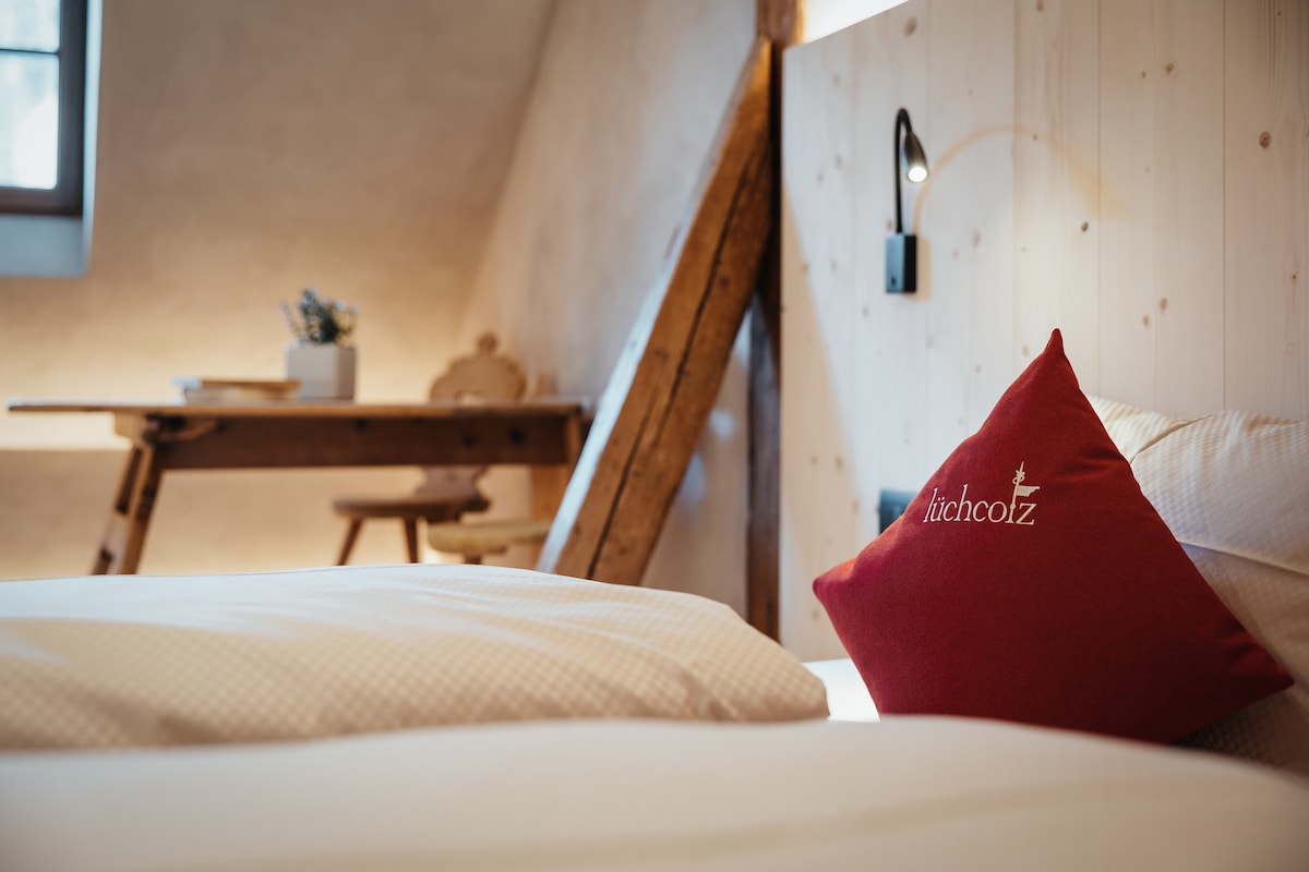 A cozy bedroom features soft bedding with a decorative red pillow embroidered with the name of the property. In the background, a wooden table and chair create a simple reading nook, complemented by exposed wooden beams and a nearby window allowing natural light.