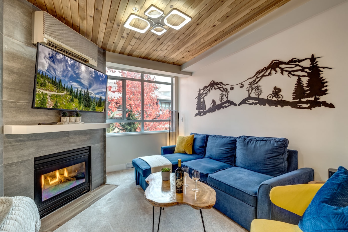 A comfortable living area features a blue sectional sofa complemented by a wooden coffee table. A mounted television displays scenic mountain imagery, and a fireplace is situated below. Large windows allow natural light to fill the space, while a nature-inspired wall art piece enhances the decor.