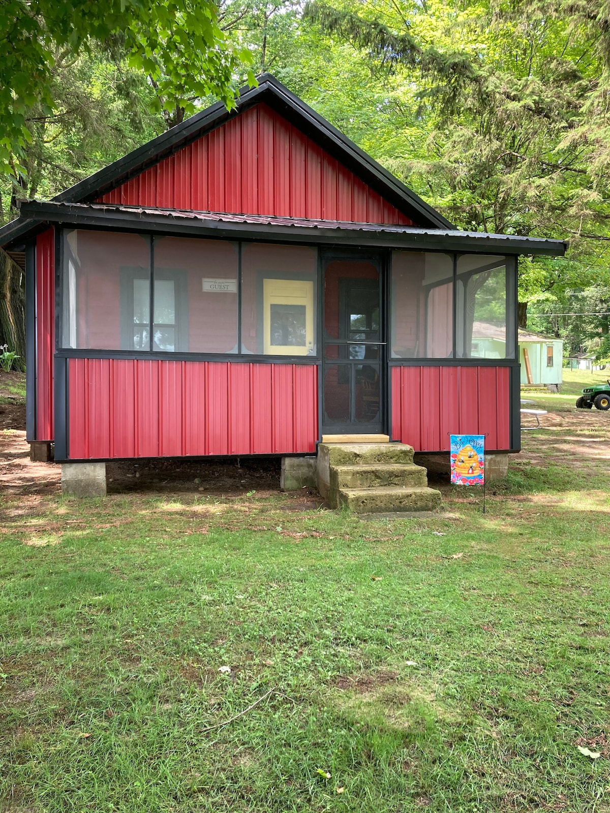 A charming red cabin is framed by lush greenery, featuring a screened-in porch that offers views of the surrounding nature. The entrance is accessible by a few stone steps, and a colorful welcome sign is positioned by the doorway.