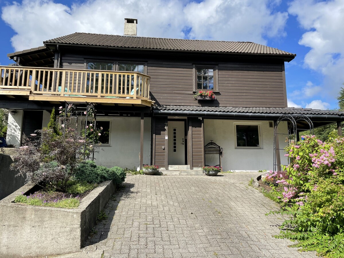 A two-story wooden house features a spacious balcony adorned with potted plants. The front entrance is framed by a landscaped area with shrubs and flowers. Bright blue skies with scattered clouds provide a serene backdrop to the welcoming exterior.