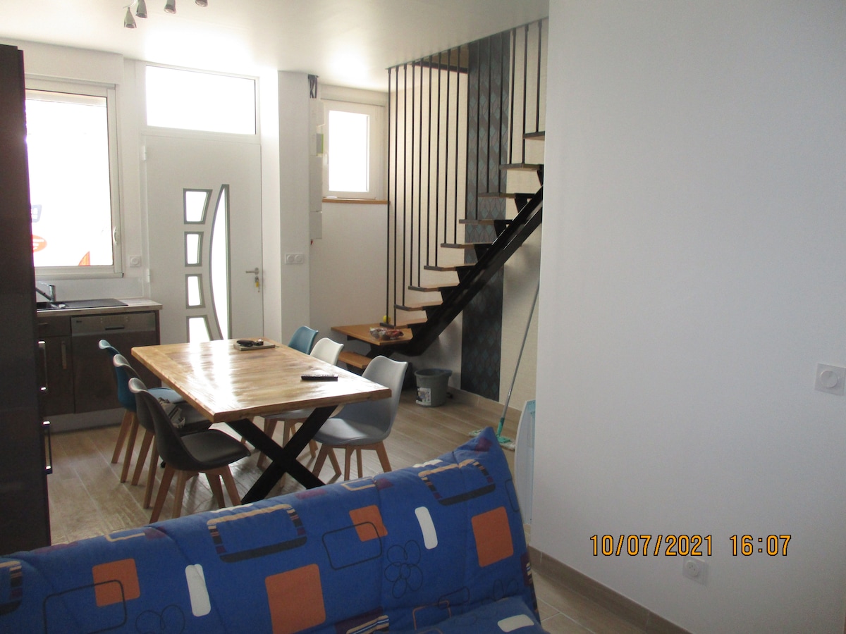 A modern dining area features a wooden table surrounded by six chairs, positioned adjacent to the kitchen. Stairs with a minimalist design ascend to the upper level, while large windows allow natural light to illuminate the space. A cozy couch is visible, adding to the inviting ambiance.