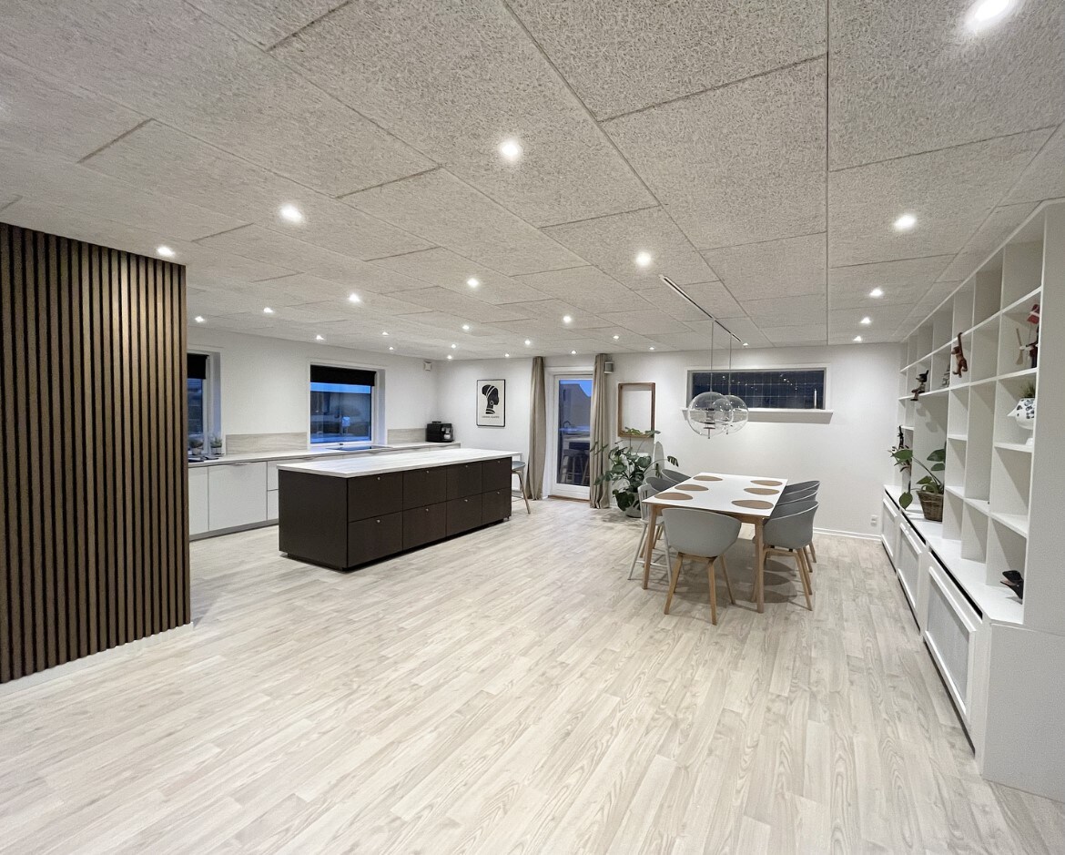 An open-plan living space features a modern dining table surrounded by several chairs. A dark kitchen island contrasts with light cabinetry and is complemented by sleek pendant lighting. Natural light fills the room through large windows, enhancing the spacious and welcoming environment.