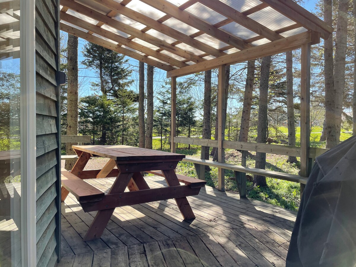 An outdoor deck area is presented, featuring a sturdy wooden picnic table surrounded by benches. A slatted roof provides partial shade, while surrounding trees contribute to a natural setting. The wooden flooring reflects a sunlit atmosphere, enhancing the peaceful outdoor experience.