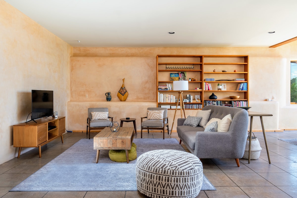 A spacious living area features a soft gray sofa and two wooden armchairs. A natural wood coffee table sits on a light area rug, while a bookshelf displays an assortment of books. A flat-screen TV rests on a wooden media console, complemented by ambient light from large windows.