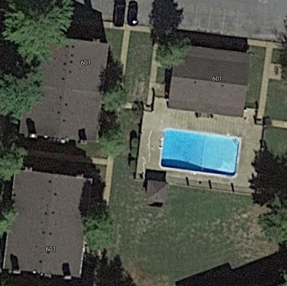 An aerial view of a community layout is displayed, featuring three identical buildings labeled '601' surrounding a centrally located swimming pool. The pool area is highlighted by a concrete patio, and green grassy spaces are visible between the buildings, offering outdoor leisure options.