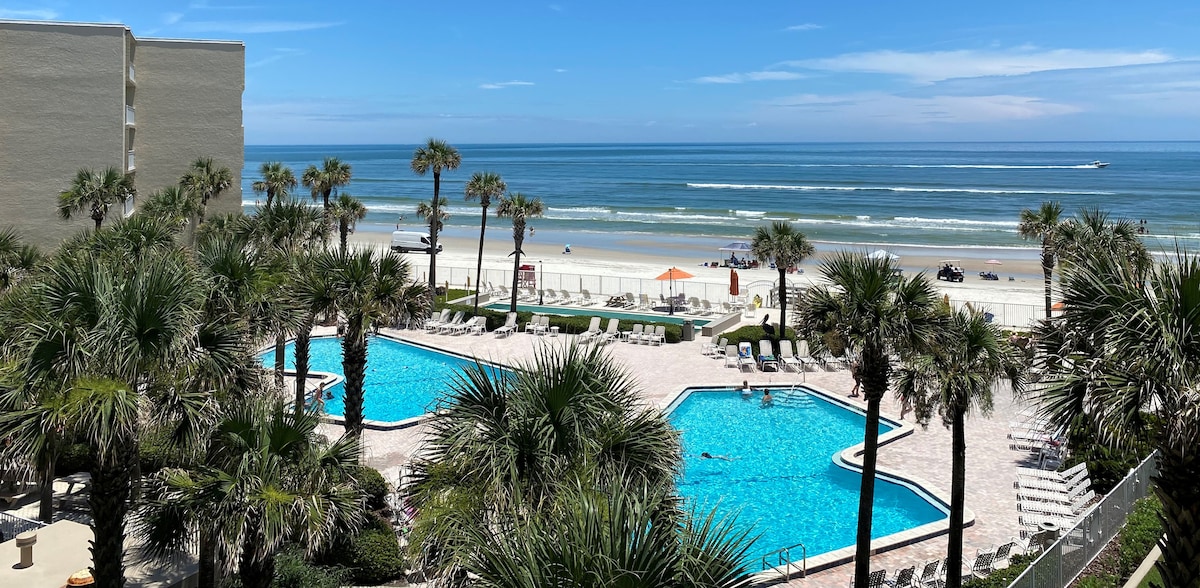 A panoramic view captures two shimmering swimming pools surrounded by tropical palm trees, with the ocean visible in the background. Beach chairs are arranged around the pools, and a sandy beach extends towards the horizon, meeting the gentle waves under a clear blue sky.