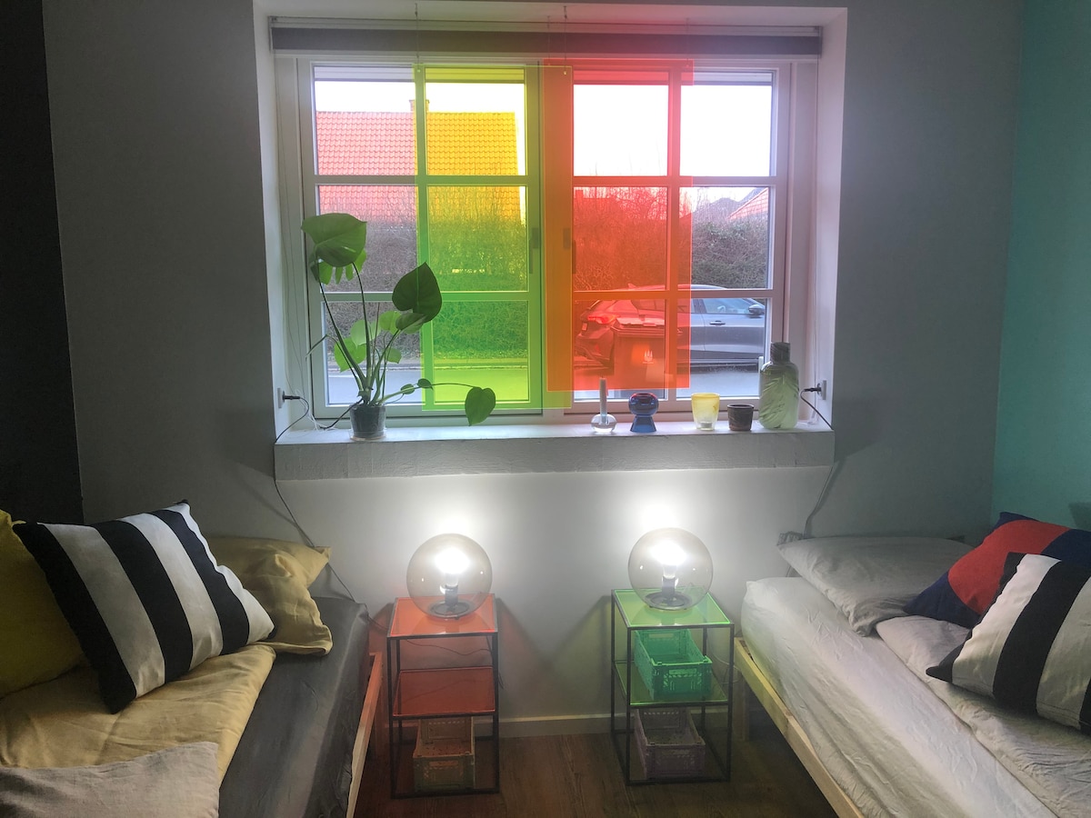 A vibrant window allows colorful light to filter into the room, casting hues on the nearby walls. Two beds are positioned on either side, accompanied by modern nightstands featuring glowing lamps. A potted plant sits on the windowsill, enhancing the lively ambiance.
