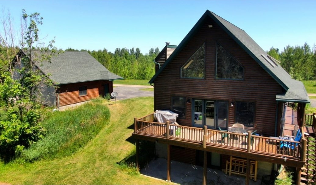 The cabin showcases a rustic exterior with a combination of wood siding and large windows. A spacious deck is visible, featuring chairs and a grill, alongside a neat lawn surrounded by greenery. A nearby separate structure can be seen in the background.