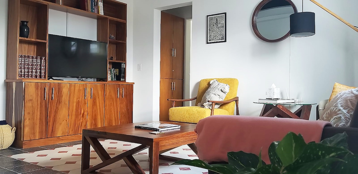 The living area is furnished with a wooden coffee table and a stylish yellow armchair. A mounted television is located on a wooden entertainment unit. A soft rug lies beneath, and a decorative plant adds a touch of greenery to the space.