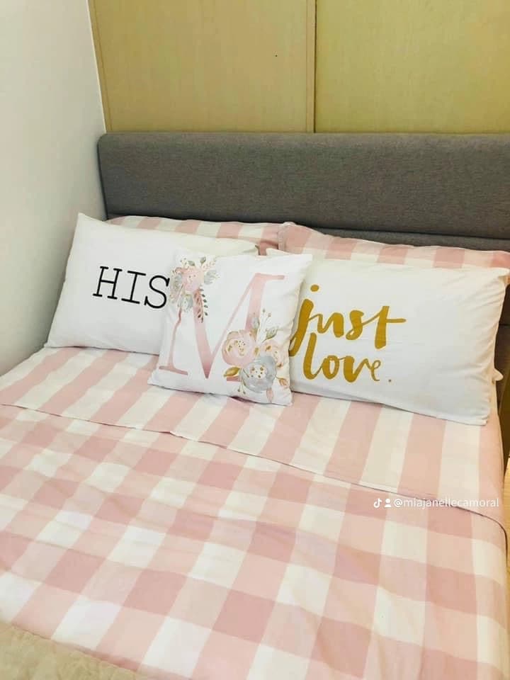A cozy sleeping area features a neatly made bed with a light pink and white checkered blanket. Decorative pillows with the words 'HIS' and 'just love' in gold lettering add a personal touch to the space.