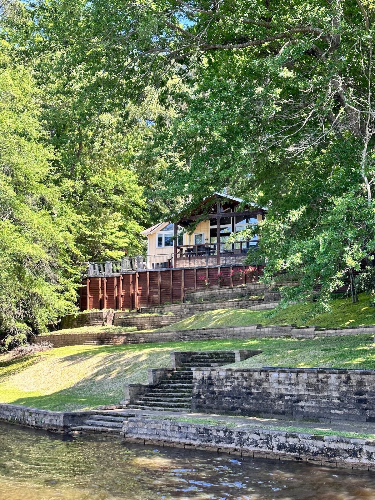 A charming cabin is elevated on a hillside, surrounded by lush greenery. Steps lead from the water's edge to the spacious deck that overlooks the lake. The wooden structure features a large porch, offering scenic views of the tranquil surroundings.