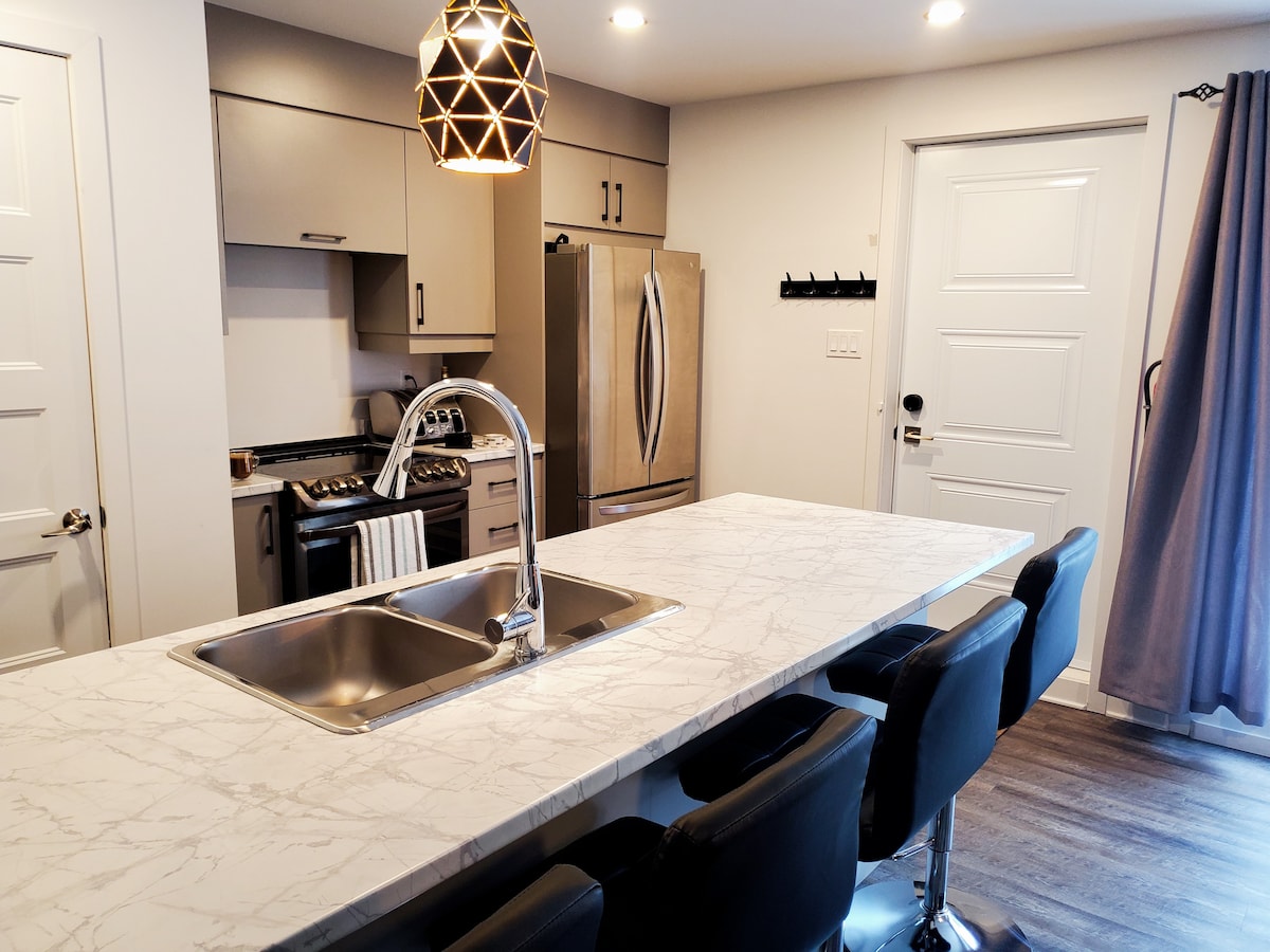 A modern kitchen is showcased, featuring a spacious marble countertop with seating for four and a sleek double sink. Stainless steel appliances, including a refrigerator, oven, and stovetop, complement the minimalist cabinetry. Natural light filters through a nearby window, enhancing the inviting ambiance.