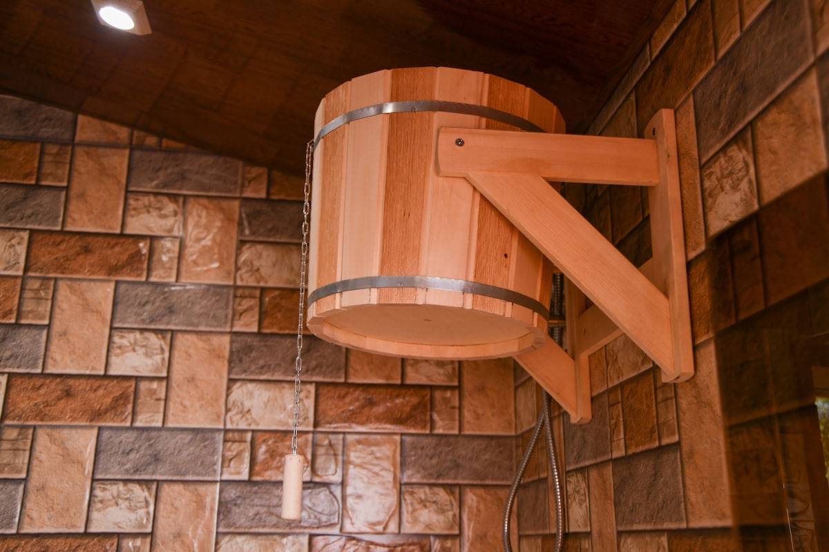 A wooden bucket is mounted on the wall, designed for a sauna experience. It features a vertical chain for easy operation and warm-toned wooden accents stand out against the textured stone tiles surrounding it.