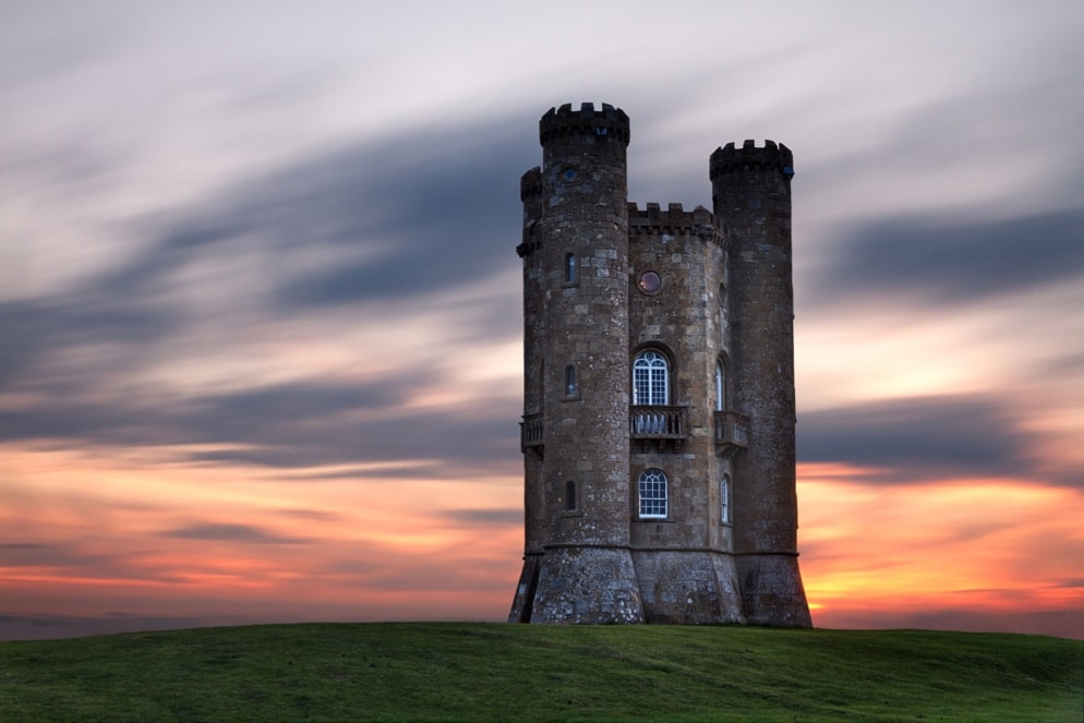 A stone tower with two rounded turrets stands prominently against a colorful sunset sky, surrounded by a gently sloping green landscape. The tower features arched windows and a decorative stone pattern, creating a striking silhouette in the fading light.