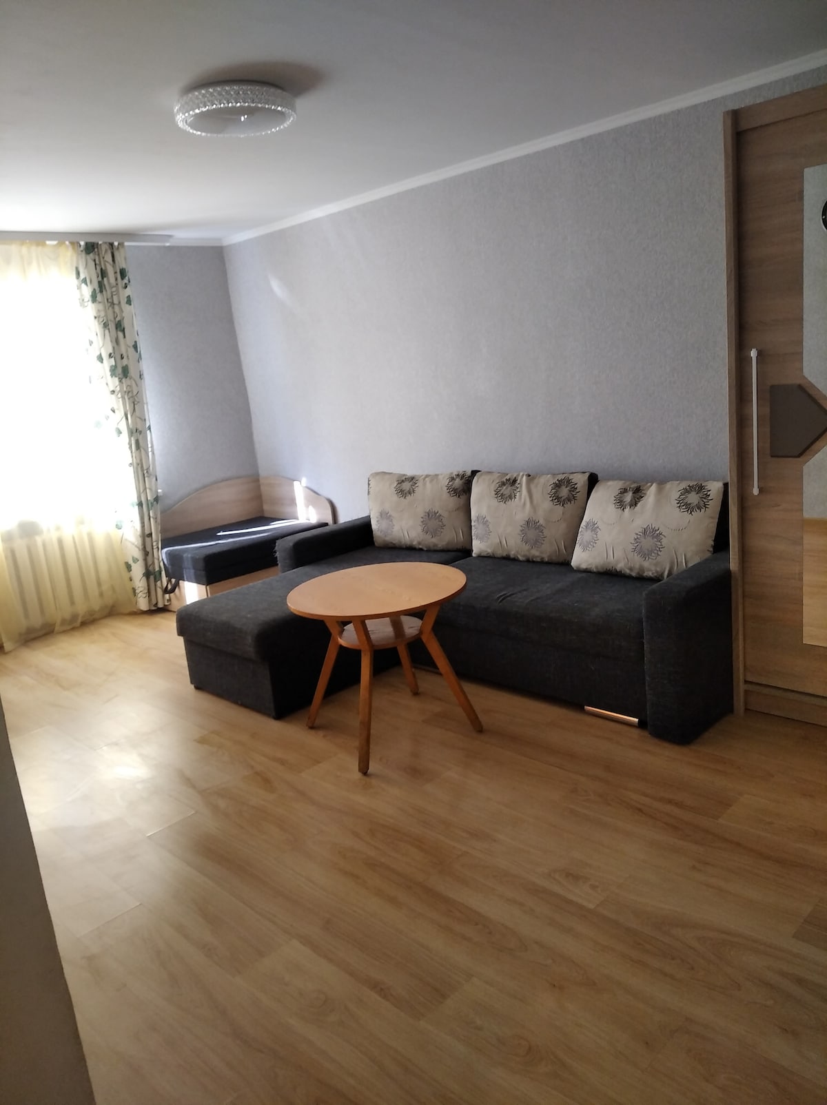 A small seating area is presented with a dark gray convertible couch adorned with patterned cushions. A round wooden table stands nearby, and natural light filters through a window, highlighting the warm wooden flooring.