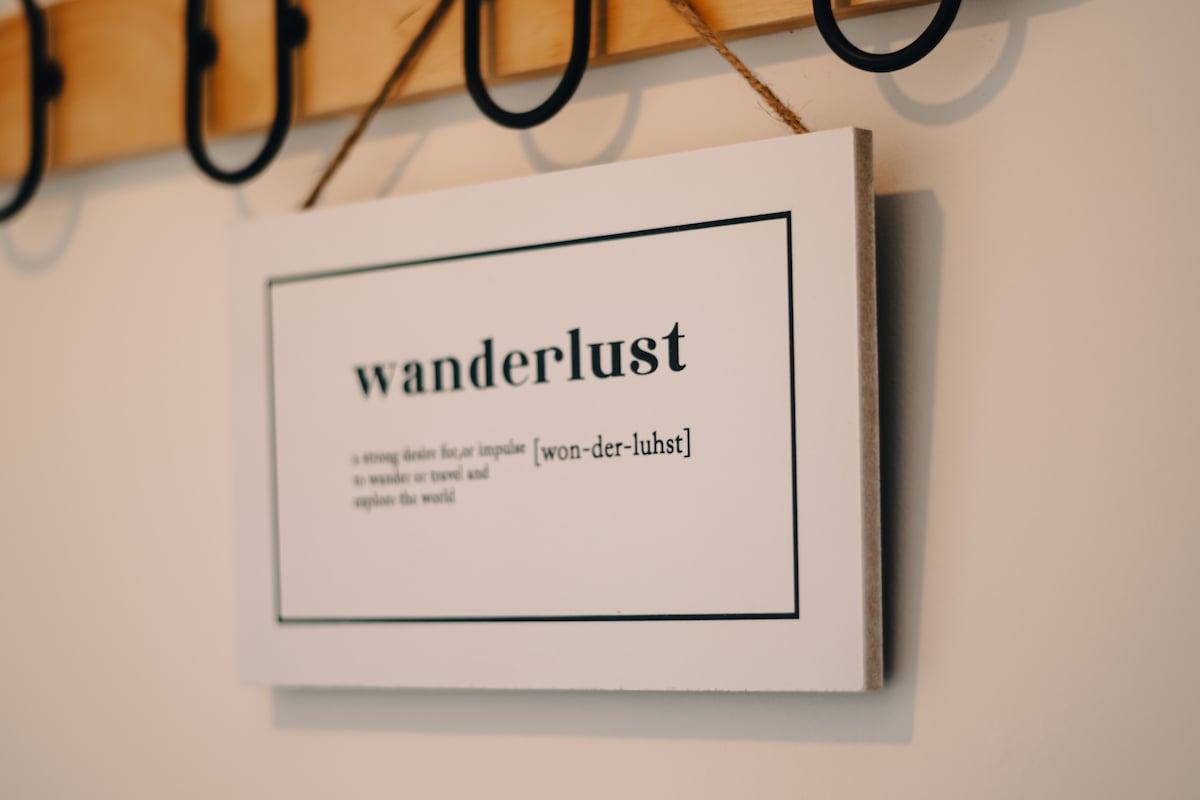 A decorative wall sign displaying the word 'wanderlust' and its definition is seen hanging on a wooden surface. The sign is framed in a simple black border, with clear, bold text emphasizing the concept of exploration.
