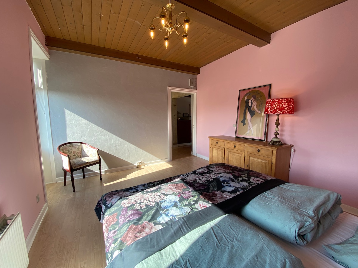 A spacious sleeping area features pale pink walls and a wooden ceiling. A large bed with floral bedding is centered in the room. A wooden dresser and a decorative lamp with a patterned shade add charm. Natural light streams through an adjacent doorway.