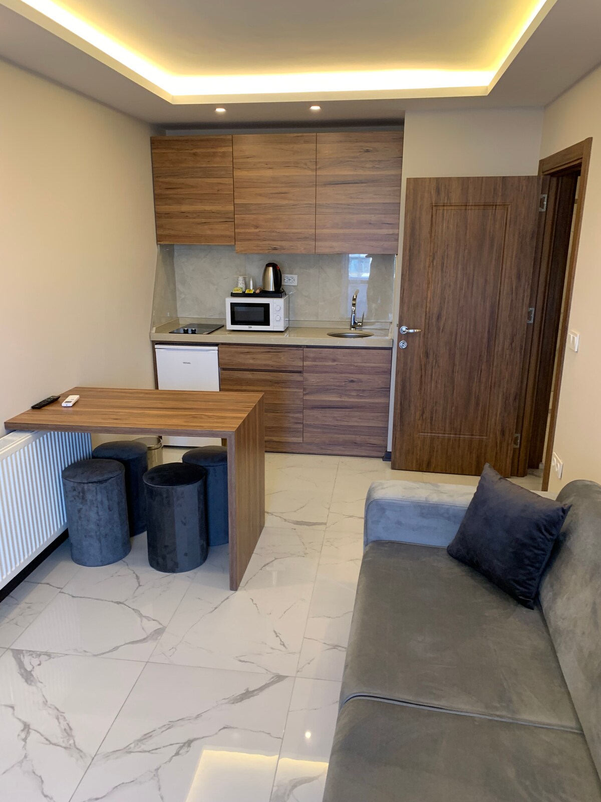 A compact kitchen area is presented, featuring wooden cabinets and a light-colored countertop. Small appliances, including a microwave and kettle, are visible. A dining table with two stools stands next to the kitchen, while a cozy seating area is arranged with a grey sofa.