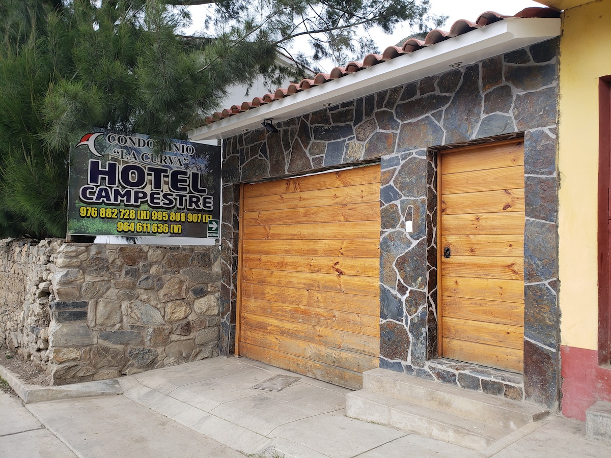 The exterior of a rustic hotel is shown, featuring a stone façade and wooden garage doors. A prominent sign displaying 'Hotel Campestre' hangs above the entrance. Surrounding pine trees add a natural element to the setting.