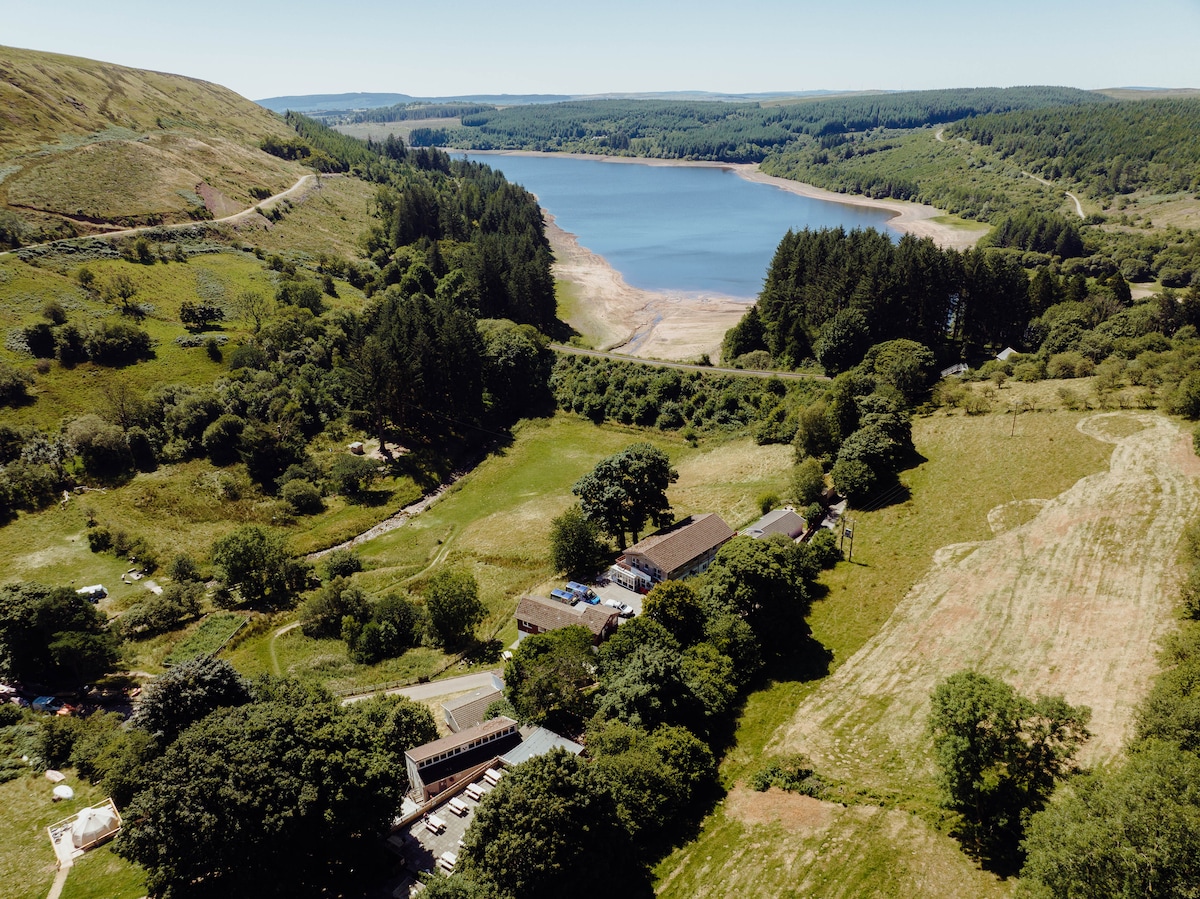 An aerial view captures the lush greenery surrounding Pontsticill Reservoir. The calm waters reflect the blue sky, bordered by rolling hills. In the foreground, a series of buildings and trees blend into the landscape, showcasing the campsite's natural setting.