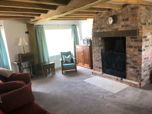 A cozy sitting room features exposed wooden beams and a warm brick fireplace. Soft seating is arranged around a central hearth, complemented by a small table and a stylish cabinet. Large windows with light blue curtains allow natural light to fill the space.