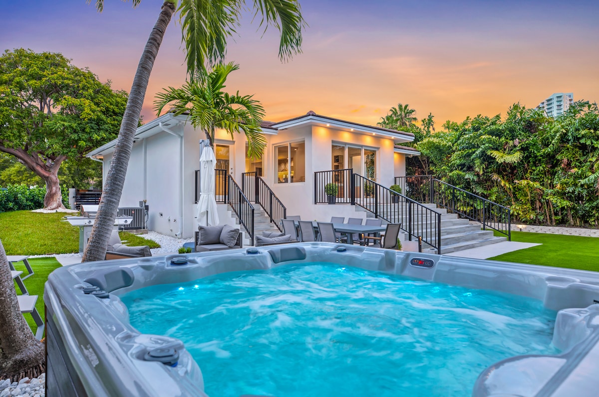 A hot tub is prominently featured in the foreground, surrounded by lush greenery and a well-manicured lawn. The modern house is visible in the background, with multiple large windows reflecting the vibrant sky at sunset.