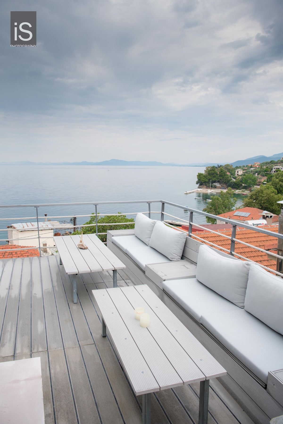 An outdoor terrace is furnished with light-colored sofas and tables, offering a space for relaxation. The area provides unobstructed views of the calm sea and nearby coastal scenery, enhancing the serene atmosphere of the setting.